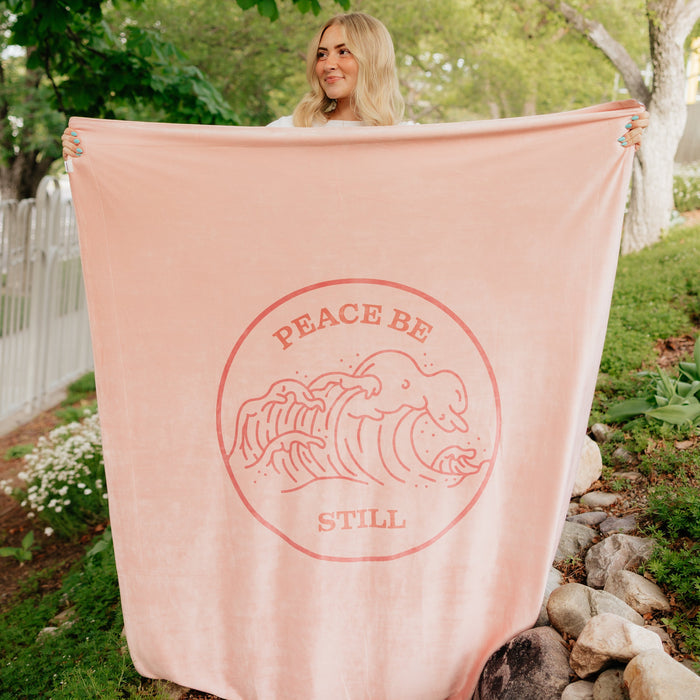 Soft blush pink Christian blanket featuring the Peace Be Still scripture design and ocean wave artwork, displayed outdoors in a peaceful garden setting