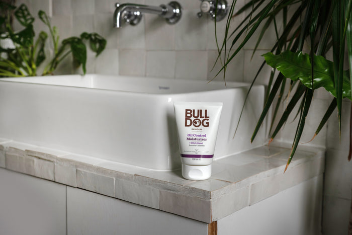 A tube of Oil Control Moisturiser rests on the rim of a white bathroom sink, surrounded by green plants and white tiled walls, highlighting this moisturiser as an ideal choice for oil-prone skin.