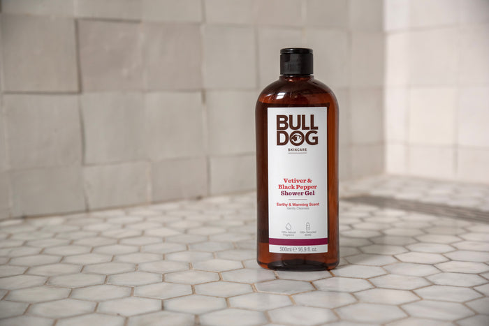 A bottle of Vetiver & Black Pepper Shower Gel, designed for men, stands on a white hexagon-tiled surface. The brown bottle has a black cap and white label, placed against a tiled wall backdrop.