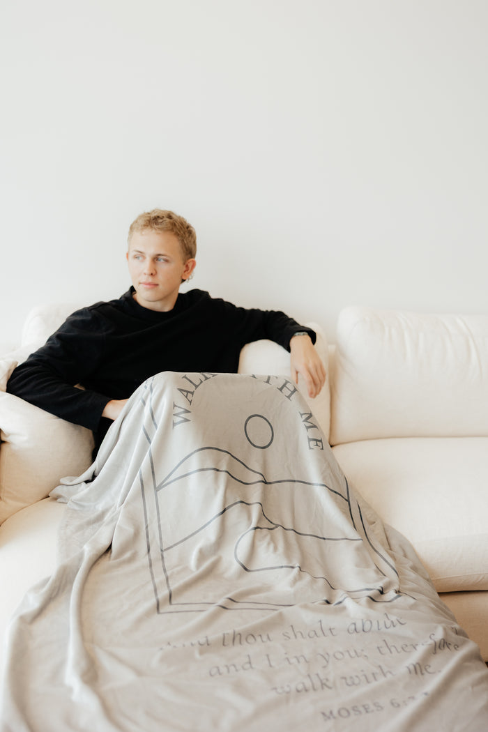 Youth sitting on a couch wrapped in a desert cream LDS youth theme blanket featuring the words "Walk With Me," faith-centered throw blanket inspired by the 2026 LDS Youth Theme and a reminder to walk with Jesus Christ.