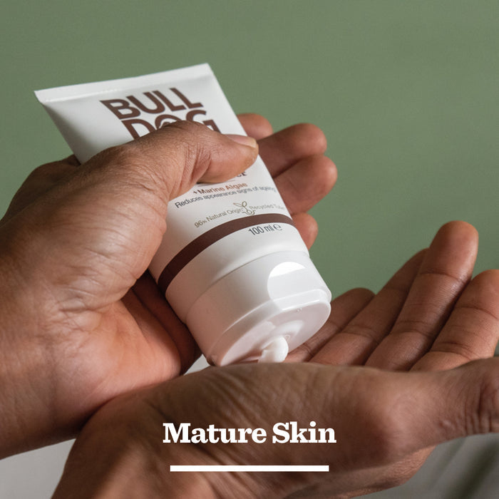 A person squeezes Age Defence Moisturiser for men onto their palm, with Mature Skin written below. The green background highlights its natural origin ingredients.