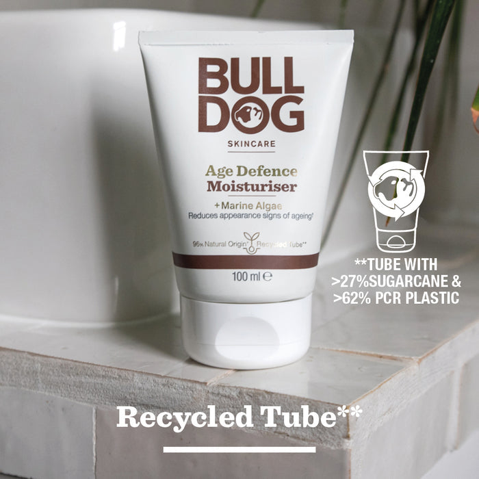 A tube of Age Defence Moisturiser for men rests on a white bathroom surface. Text states it’s made with over 27% sugarcane, 62% PCR plastic, uses natural origin ingredients, and is labeled as a Recycled Tube.
