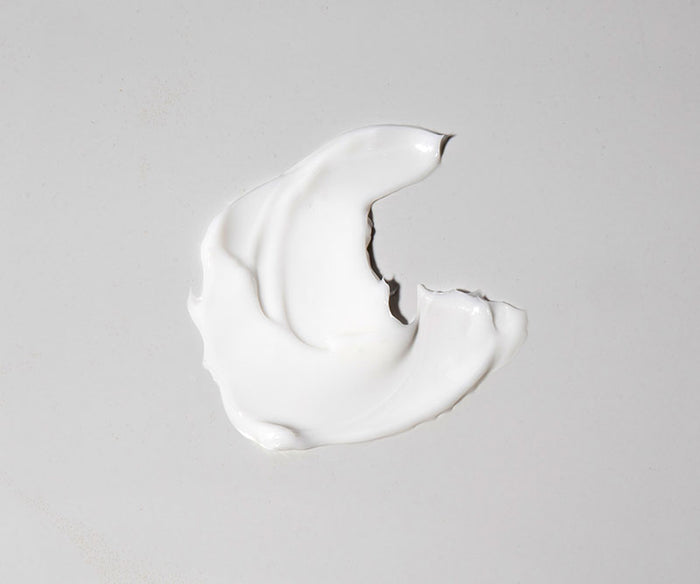 A dollop of Age Defence Moisturiser, enriched with naturally derived ingredients, is smeared in a crescent shape on a smooth, light gray surface.