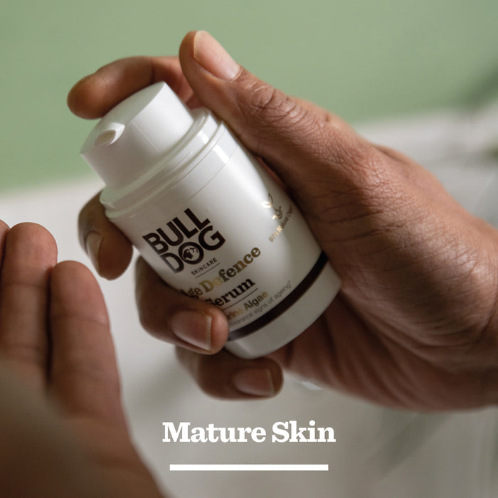 A person holds a bottle of Age Defence Serum for mature skin, rich in natural origin ingredients, about to dispense some into their palm. The softly blurred background features green and white tones.