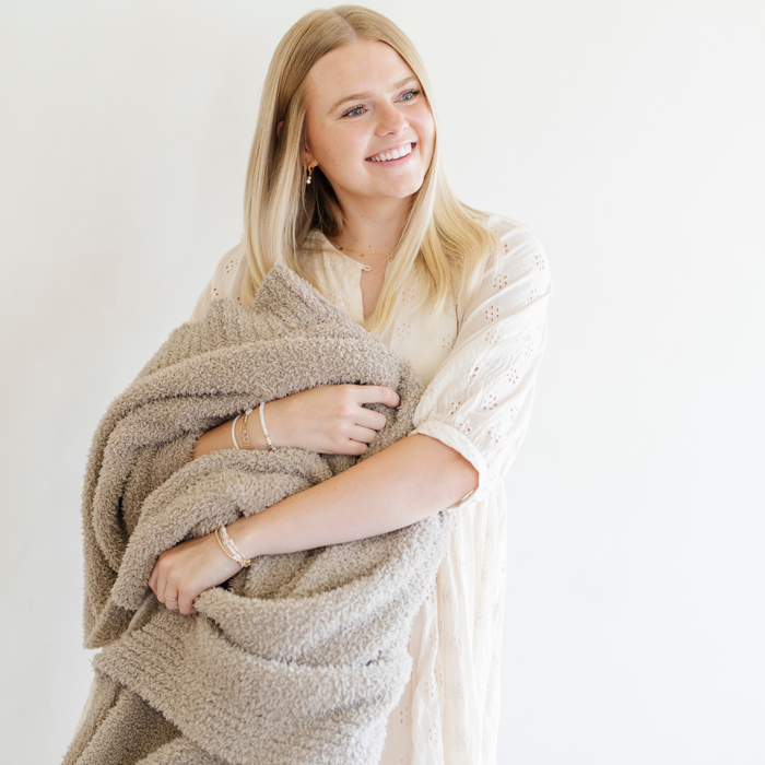 A woman smiles and holds a Bamboni® blanket by Saranoni. This ultra-soft blanket is a taupe blanket and made from a premium blend of bamboo rayon and poly microfiber, offering a lightweight, breathable, and fluffy feel. Perfect as a throw blanket, luxury blanket, plush throw, or soft blanket, it adds warmth and elegance to any living room, bedroom, or home décor setting. Designed for ultimate comfort, this buttery-soft Saranoni blanket is ideal for gifting. Enjoy this cozy blanket for years to c