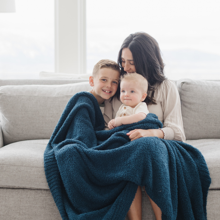 A mother and her kids sit with a Bamboni® blanket by Saranoni. This ultra-soft blanket is a blue blanket and made from a premium blend of bamboo rayon and poly microfiber, offering a lightweight, breathable, and fluffy feel. Perfect as a throw blanket, luxury blanket, plush throw, or soft blanket, it adds warmth and elegance to any living room, bedroom, or home décor setting. Designed for ultimate comfort, this buttery-soft Saranoni blanket is ideal for gifting. Enjoy this cozy blanket for years