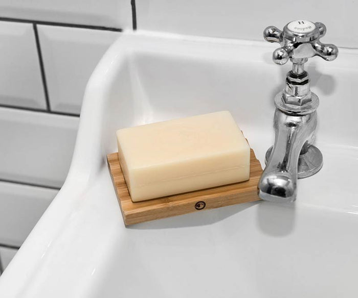 A rectangular bar of soap rests on a Bamboo Soap Dish at the edge of a white sink near a chrome faucet, with white tile and black grout behind—perfect for extending soap life and keeping grooming essentials organized.