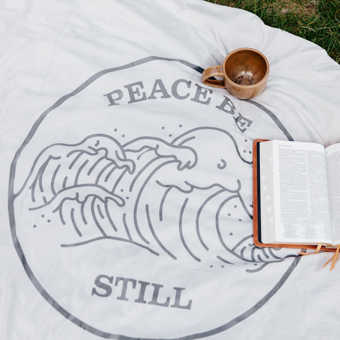 Peace Be Still blanket displayed outdoors with an open Bible and ceramic mug, featuring a calming wave design inspired by scripture.