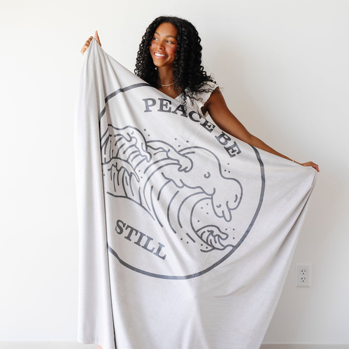 Woman holding a Christian blanket featuring the Peace Be Still scripture design and ocean wave artwork, displayed against a white background.