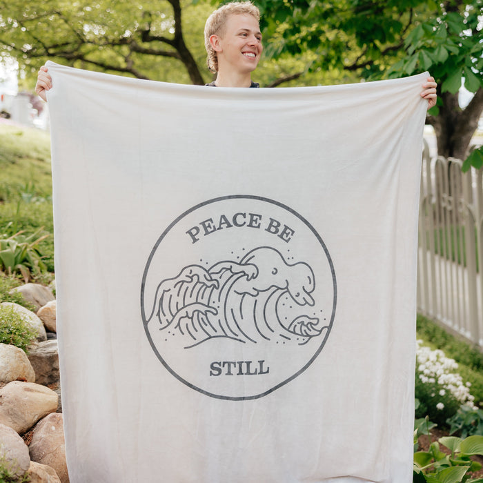 Person holding a Christian blanket featuring the Peace Be Still scripture design and ocean wave artwork outdoors.
