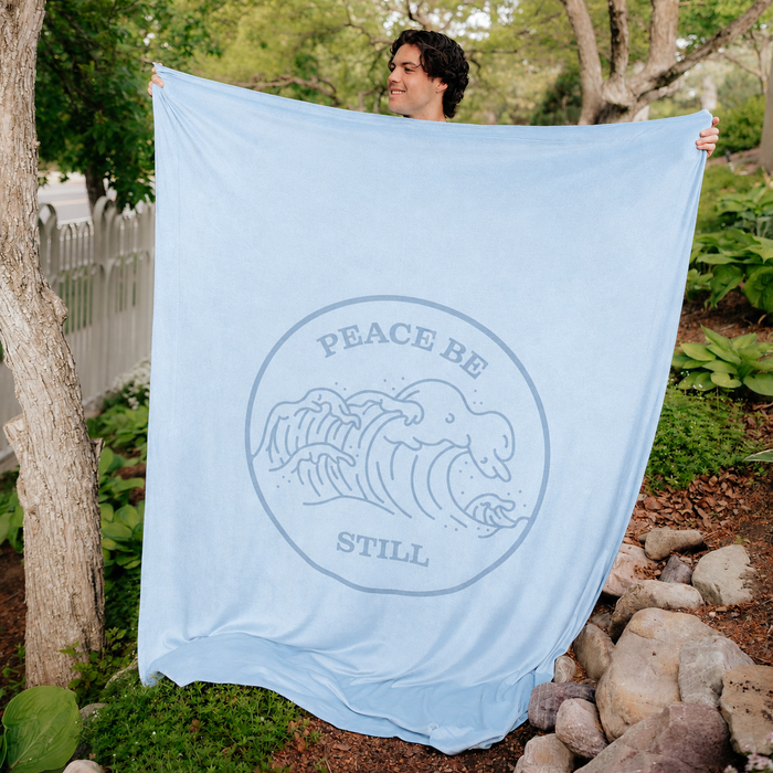 Christian blanket featuring the Peace Be Still scripture design and ocean wave artwork, displayed outdoors in a peaceful garden setting.