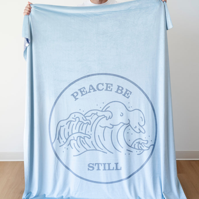 Person holding a Christian blanket featuring the Peace Be Still scripture design and ocean wave artwork, displayed on a soft blue blanket.
