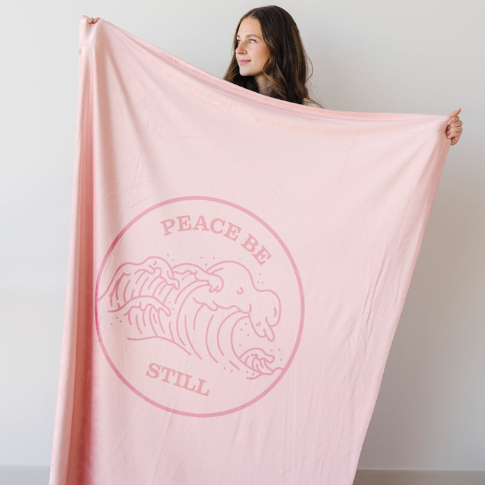 Woman holding a blush pink Christian blanket featuring the Peace Be Still scripture design and ocean wave artwork.
