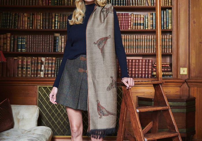 A woman modelling Dubarry women's Birchdale Tweed Taupe Stole draped over her shoulder.  She is also wearing a navy knitted jumper and a tweed skirt.