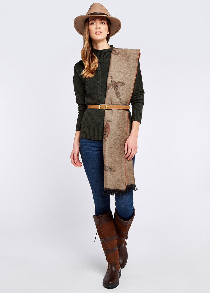 A woman modelling Dubarry women's Birchdale Tweed Taupe Stole draped over her shoulder.  She is also wearing a belt around her waist a green knitted jumper, denim honeysuckle jeans and brown country boots.