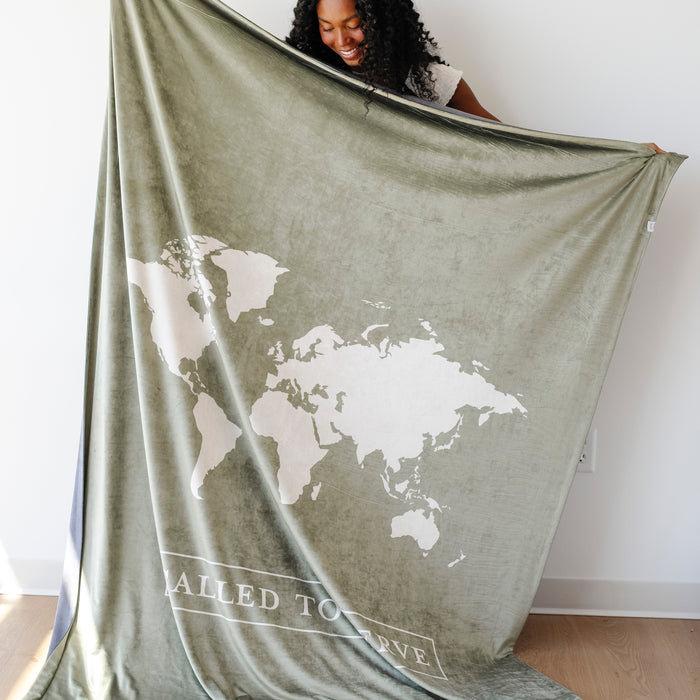 Woman holding a cypress green LDS missionary blanket featuring a world map design and the words "Called to Serve," meaningful missionary gift blanket celebrating faith, service, and missionary journeys around the world.