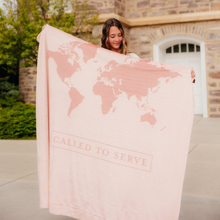 Woman holding a blush pink LDS missionary blanket featuring a world map design and the words "Called to Serve," meaningful missionary gift blanket celebrating faith, service, and missionary journeys around the world.