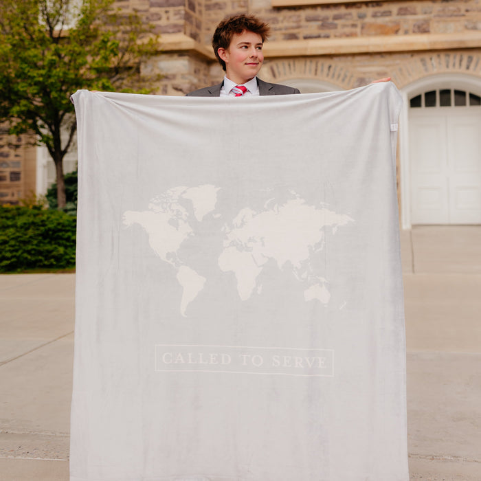 Young missionary holding a shell cream LDS missionary blanket featuring a world map design and the words "Called to Serve," meaningful missionary gift blanket celebrating faith, service, and missionary journeys around the world.