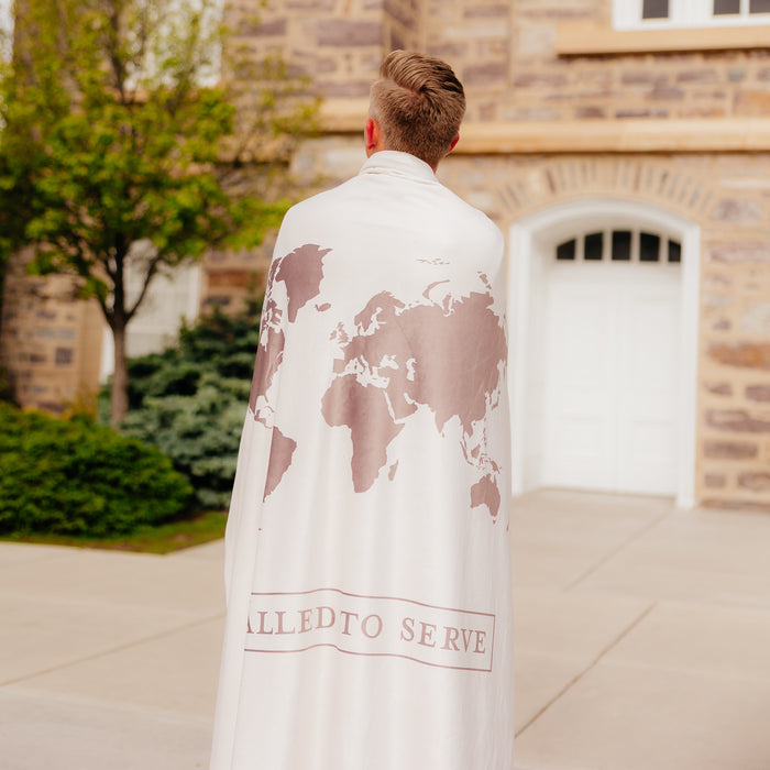 Missionary standing outdoors wrapped in a shell cream LDS missionary blanket featuring a world map design and the words "Called to Serve," meaningful missionary gift blanket celebrating faith, service, and missionary journeys around the world.