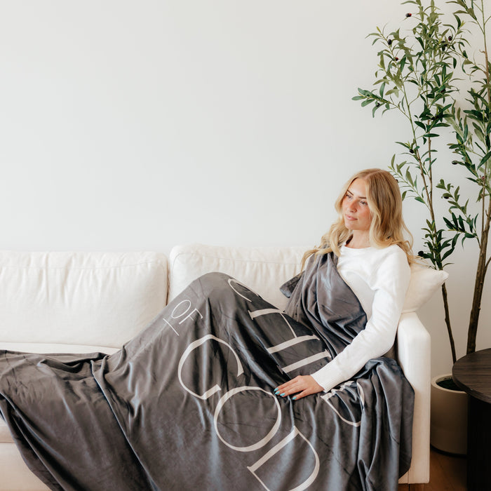 Woman relaxing on a couch wrapped in a dark obsidian Christian blanket featuring the words "Child of God," soft faith-inspired throw blanket for comfort and encouragement.