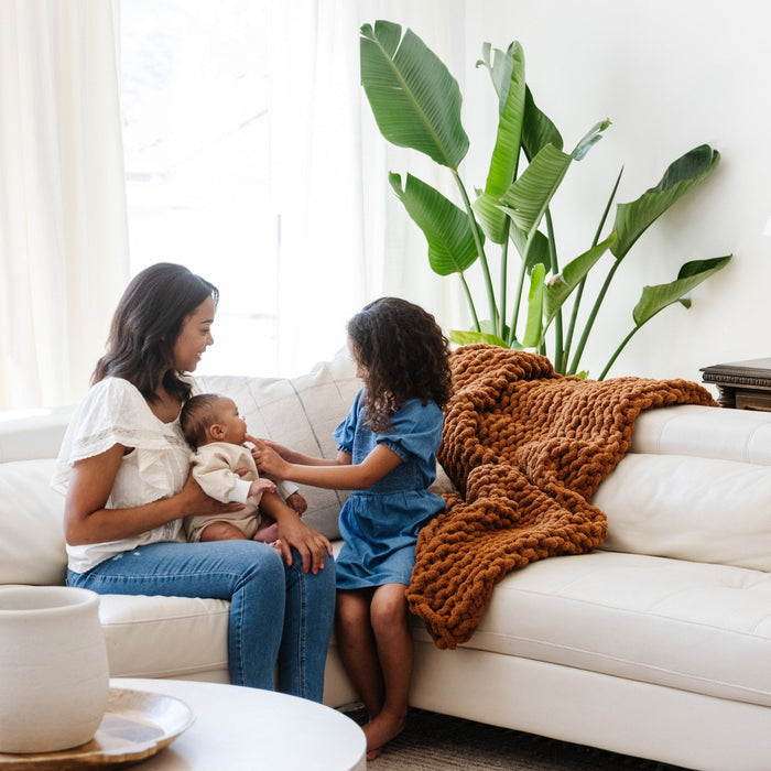 A mother and her children sit by a soft, ginger colored Saranoni chunky knit throw blanket. The large knit Saranoni blanket, is made from 100% poly microfiber and drapes beautifully. This luxury throw blanket is both trendy and durable, making it the perfect addition to any space. Ideal for home decor enthusiasts and those seeking a cuddly, soft blanket, this chunky knit blanket combines style and comfort. Keywords: chunky knit throw, soft blanket, knit blanket, large knit blanket, luxury throw
