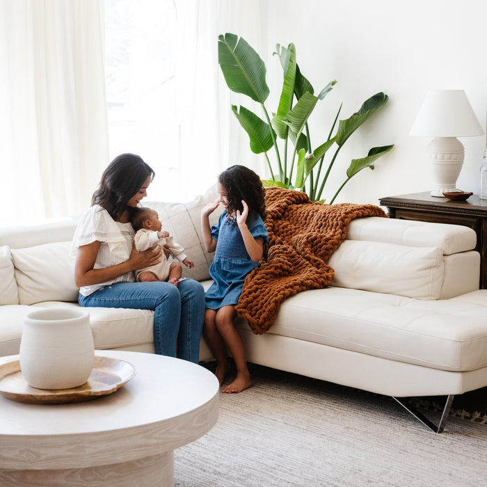 A mother and her children sit by a soft, ginger colored Saranoni chunky knit throw blanket. The large knit Saranoni blanket, is made from 100% poly microfiber and drapes beautifully. This luxury throw blanket is both trendy and durable, making it the perfect addition to any space. Ideal for home decor enthusiasts and those seeking a cuddly, soft blanket, this chunky knit blanket combines style and comfort. Keywords: chunky knit throw, soft blanket, knit blanket, large knit blanket, luxury throw