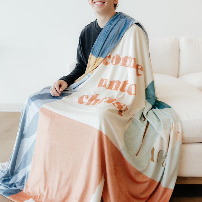 Young man sitting on a couch wrapped in a Come Unto Christ LDS blanket featuring colorful faith-centered artwork and the words "Come Unto Christ," meaningful blanket for LDS youth, missionaries, and Christian homes.