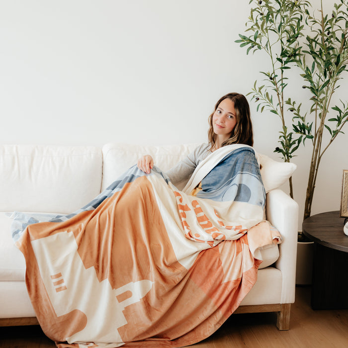 Young woman sitting on a couch wrapped in a Come Unto Christ LDS blanket featuring colorful faith-centered artwork and the words "Come Unto Christ," meaningful blanket for LDS youth, girls camp, youth conference, and Christian homes.