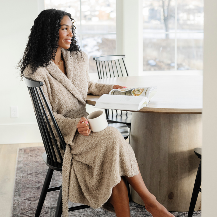 A woman is sitting at a table and wearing a Saranoni Bamboni® Robe in Taupe. Crafted from our signature ultra-soft and breathable Bamboni® fabric, this women’s plush robe wraps you in warmth while remaining lightweight. Featuring a classic shawl collar, deep pockets, and an adjustable tie waist, it's perfect for lounging, self-care mornings, and unwinding. Elevate your relaxation with the best soft robe for women, designed for everyday luxury. The soft robe is a luxury robe and a Saranoni robe.