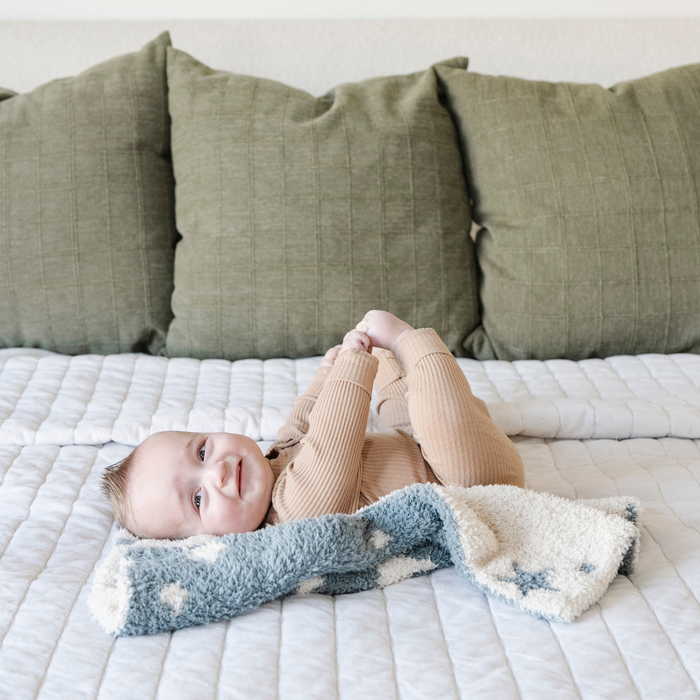 A smiling baby boy with a double-layer Bamboni® baby blanket. The plush, breathable blanket features a two-tone blue and cream star design, offering ultimate softness and warmth. Made from Saranoni’s signature stretchy, cloud-like Bamboni® fabric, this luxury baby blanket provides comfort for naps, tummy time, and on-the-go snuggles. The perfect blend of warmth and breathability, it’s a must-have for parents seeking high-quality, ultra-soft blankets. The soft blanket is a mini blanket and Sarano
