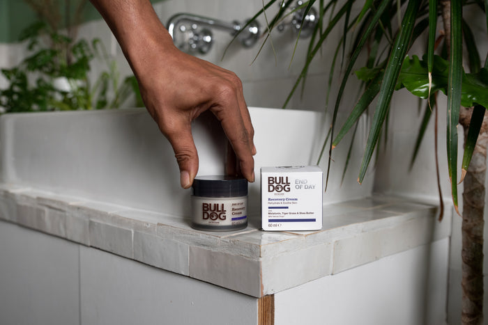 A hand reaches for a jar of End of Day Recovery Cream, placed on a bathroom counter beside its box, with green plants and a white sink in the background.