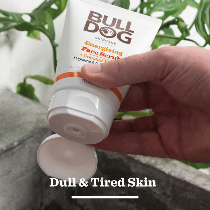 A hand squeezes a tube of Energising Face Scrub for men, made with natural origin ingredients, against green plants and concrete. Text overlay: “Dull & Tired Skin.”.