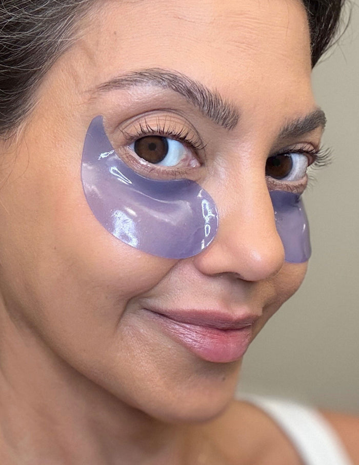 Close-up of a woman wearing purple under-eye gel patches and smiling slightly
