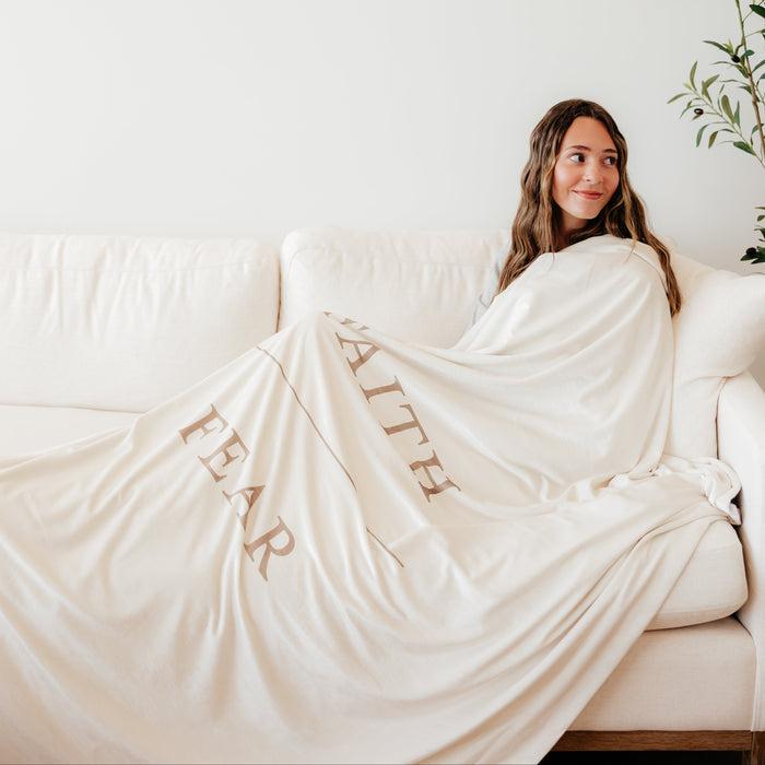 Woman wrapped in a shell cream Christian blanket featuring the words "Faith Over Fear," soft faith-inspired throw blanket offering comfort, encouragement, and a reminder to trust in God through life's challenges.