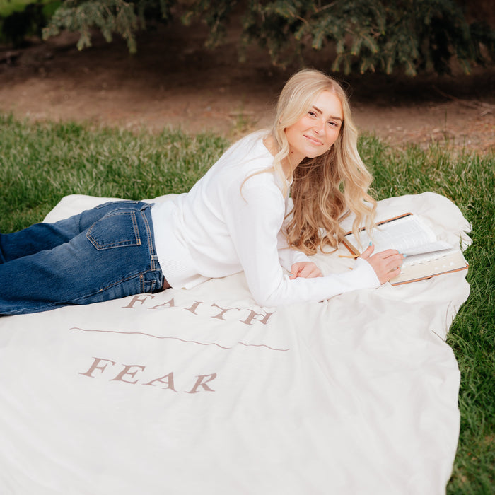 Young woman reading scriptures while lying on a shell cream Christian blanket featuring the words "Faith Over Fear," faith-inspired throw blanket offering comfort, encouragement, and a reminder to trust God through life's challenges.