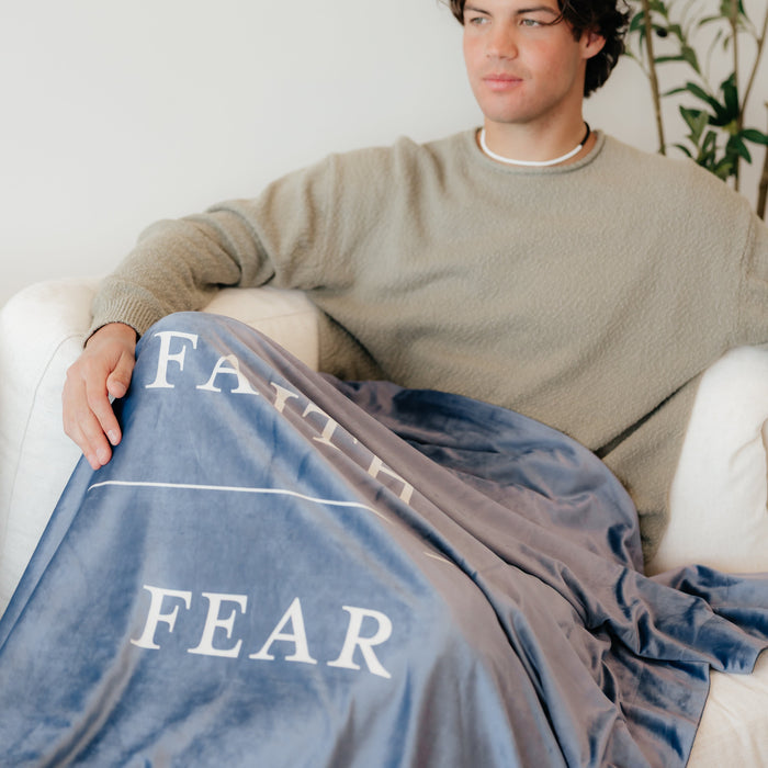 Young man sitting on a couch wrapped in a harbor blue Christian blanket featuring the words "Faith Over Fear," faith-inspired throw blanket offering comfort, encouragement, and a reminder to trust God through life's challenges.