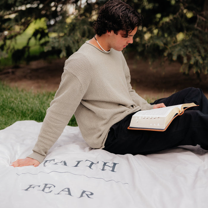 Young man reading scriptures while sitting on a shell cream Christian blanket featuring the words "Faith Over Fear," faith-inspired throw blanket offering comfort, encouragement, and a reminder to trust God through life's challenges.