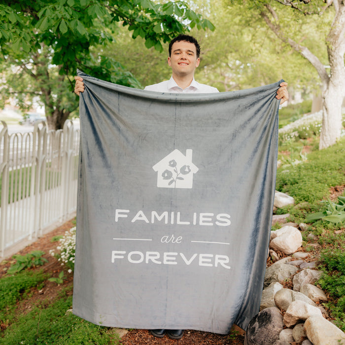 Person holding a dark obsidian LDS blanket featuring the words "Families Are Forever" and a home design outdoors, soft faith-inspired throw blanket celebrating eternal families and lasting connections.