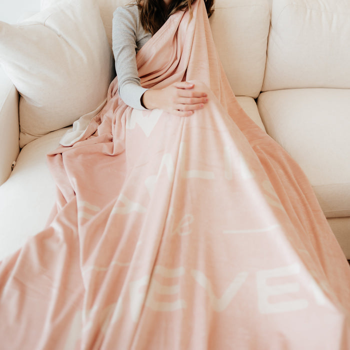 Woman relaxing on a couch wrapped in a soft peony pink LDS blanket featuring the words "Families Are Forever," faith-inspired throw blanket celebrating eternal families and meaningful connections.