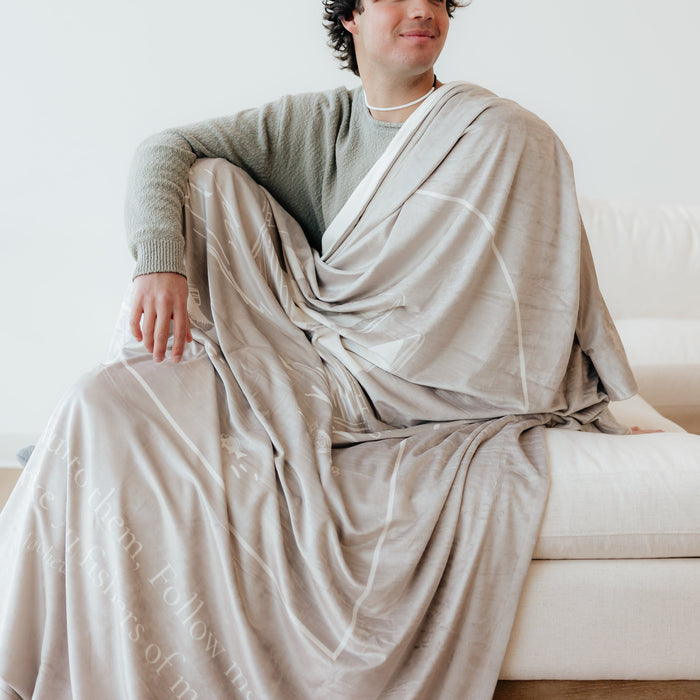 Man wrapped in a cobblestone Fishers of Men Christian blanket featuring Matthew 4:19 scripture while seated on a white couch