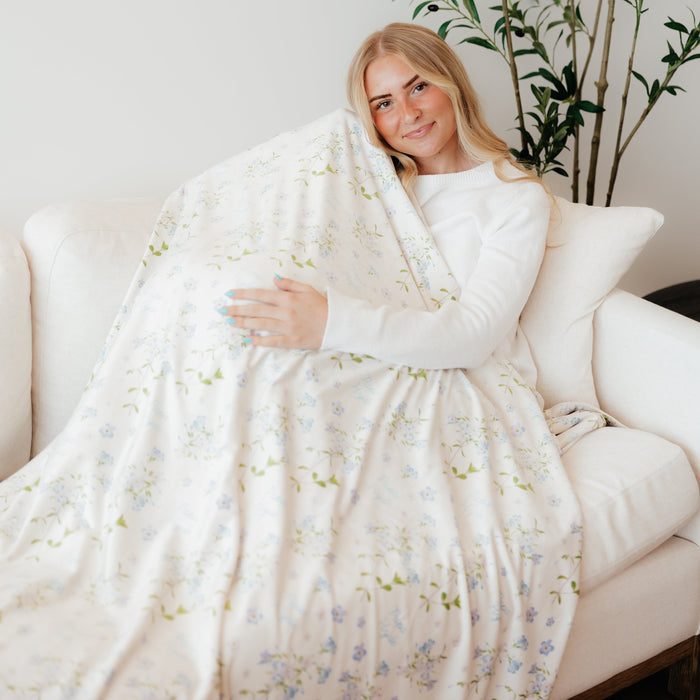Woman sitting on a couch wrapped in a personalized Forget Me Not sympathy blanket featuring delicate floral artwork and custom name details, meaningful memorial gift blanket offering comfort, remembrance, and support during times of loss.