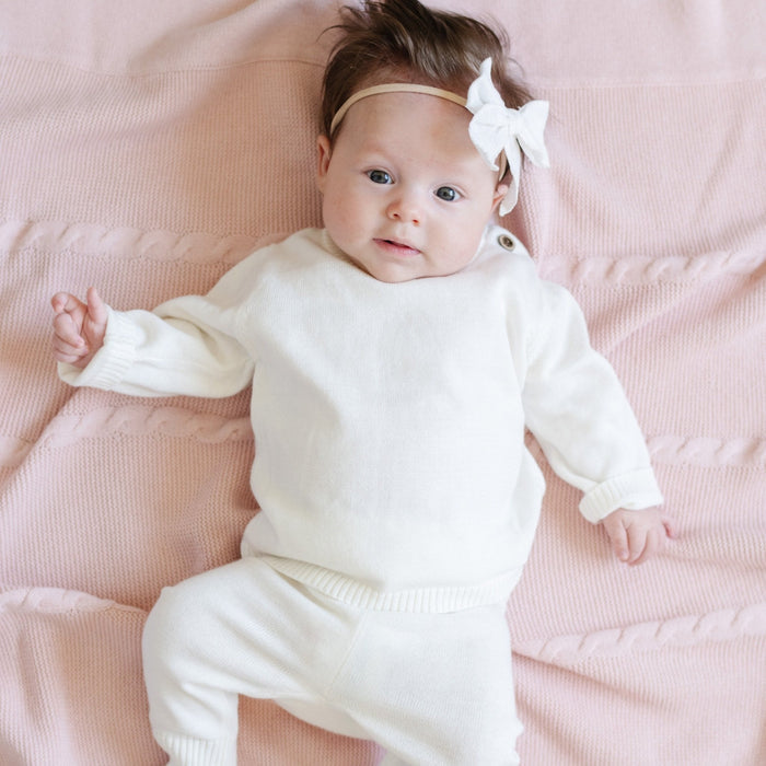 A baby girl lays on a Saranoni Heirloom Knit Blanket. She is wearing an all-white, 100% cotton baby outfit that combines style and comfort. The Saranoni cloud-soft cotton set features a breathable waistband, loosely fitted sleeves, and elevated button details, making it perfect for everyday wear or special occasions. This heirloom-quality baby clothing offers timeless style and ultimate comfort. Ideal for parents searching for luxury baby clothes, soft cotton baby sets, or high-quality infant ou