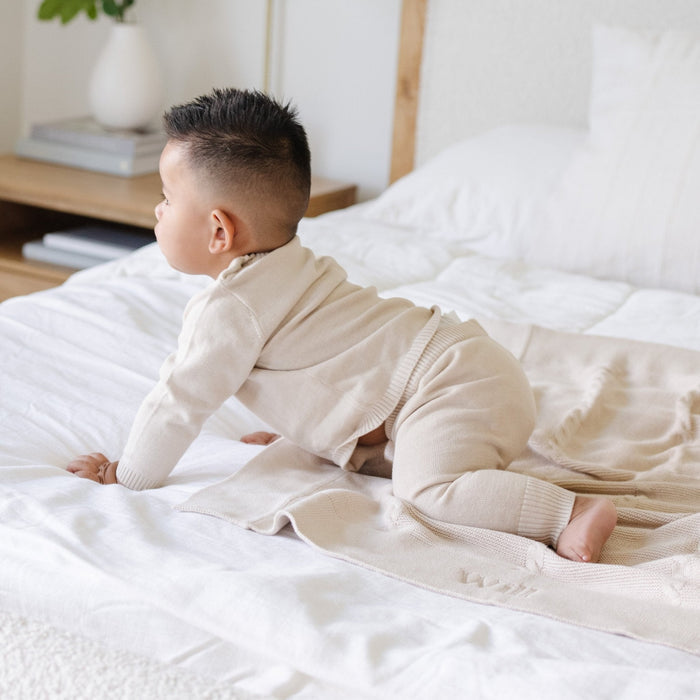 A baby boy crawls on a Saranoni Heirloom Knit Blanket. He is wearing a light tan, 100% cotton baby outfit that combines style and comfort. The Saranoni cloud-soft cotton set features a breathable waistband, loosely fitted sleeves, and elevated button details, making it perfect for everyday wear or special occasions. This heirloom-quality baby clothing offers timeless style and ultimate comfort. Ideal for parents searching for luxury baby clothes, soft cotton baby sets, or high-quality infant out