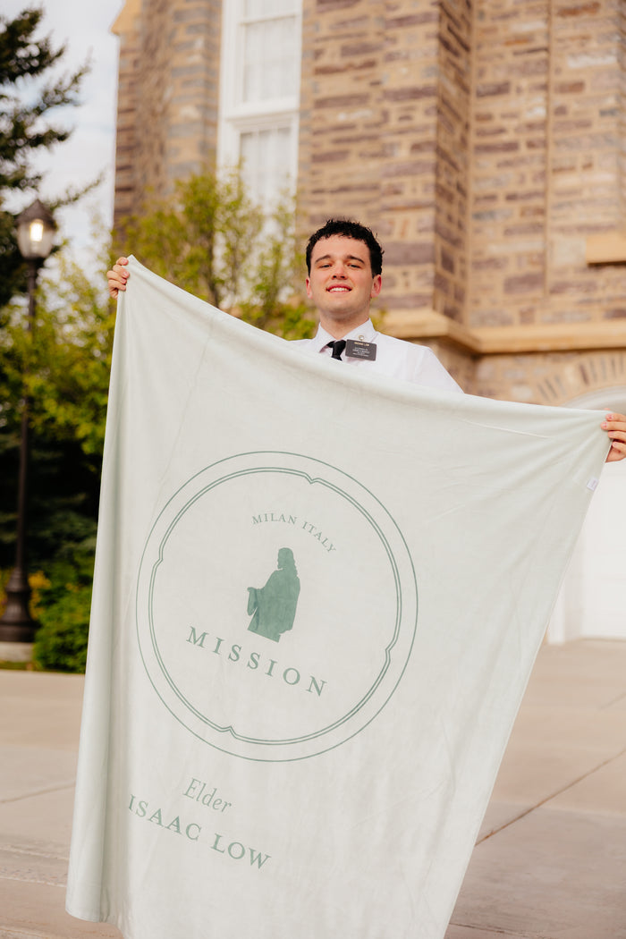 Missionary holding an In His Service LDS missionary blanket featuring personalized missionary details and faith-centered artwork outside a church building.