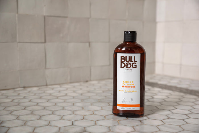 A bottle of Lemon & Bergamot Shower Gel sits on white hexagonal tiles with beige squares behind, showcasing this mens skincare essential and its refreshing, natural fragrance.