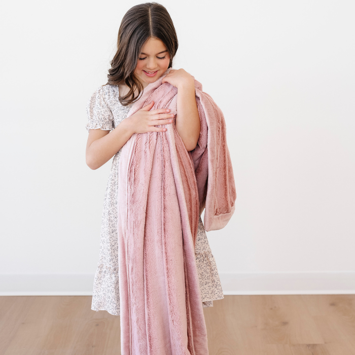 A little girl is smiling and holding a kid's blanket that is a lush pink blanket. This soft blanket is a toddler blanket and a Saranoni blanket. This blanket is a great toddler gift or addition to a bedroom.