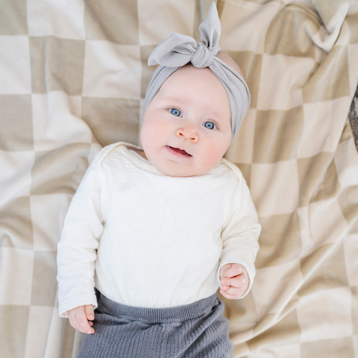 A baby girl lays on a neutral checkered minky stretch blanket. The soft blanket is a luxury blanket and a Saranoni blanket. This cozy blanket is also a receiving blanket and is a cute and cozy baby blanket. This stretchy blanket is a great nursery item.