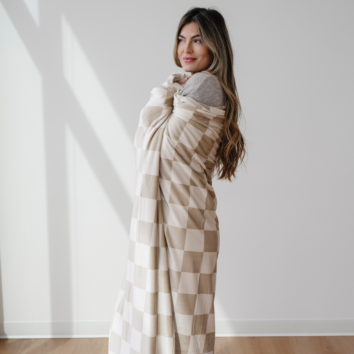 A woman is wrapped in a tan and cream checkered minky stretch blanket. This soft blanket is a Saranoni luxury blanket, and it is very stretchy, durable, and breathable. This extra large throw blanket is a very cozy blanket as well.