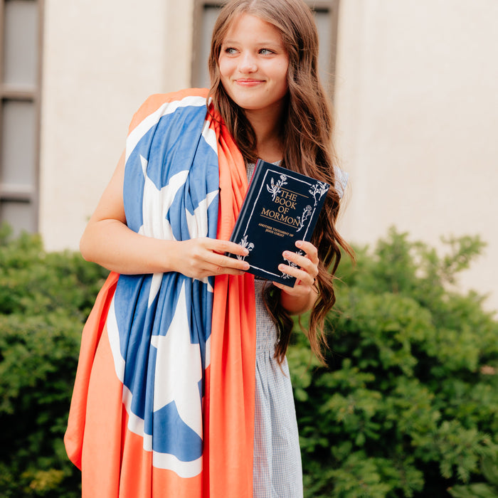 Young woman wrapped in a personalized mission flag blanket while holding a Book of Mormon, custom LDS missionary blanket celebrating faith, missionary service, and meaningful gifts.