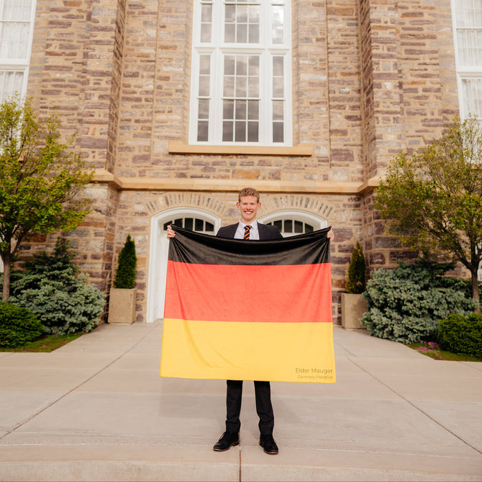 Missionary holding a personalized Germany mission flag blanket in front of a church building, custom LDS missionary keepsake blanket celebrating missionary service, mission calls, and faith.